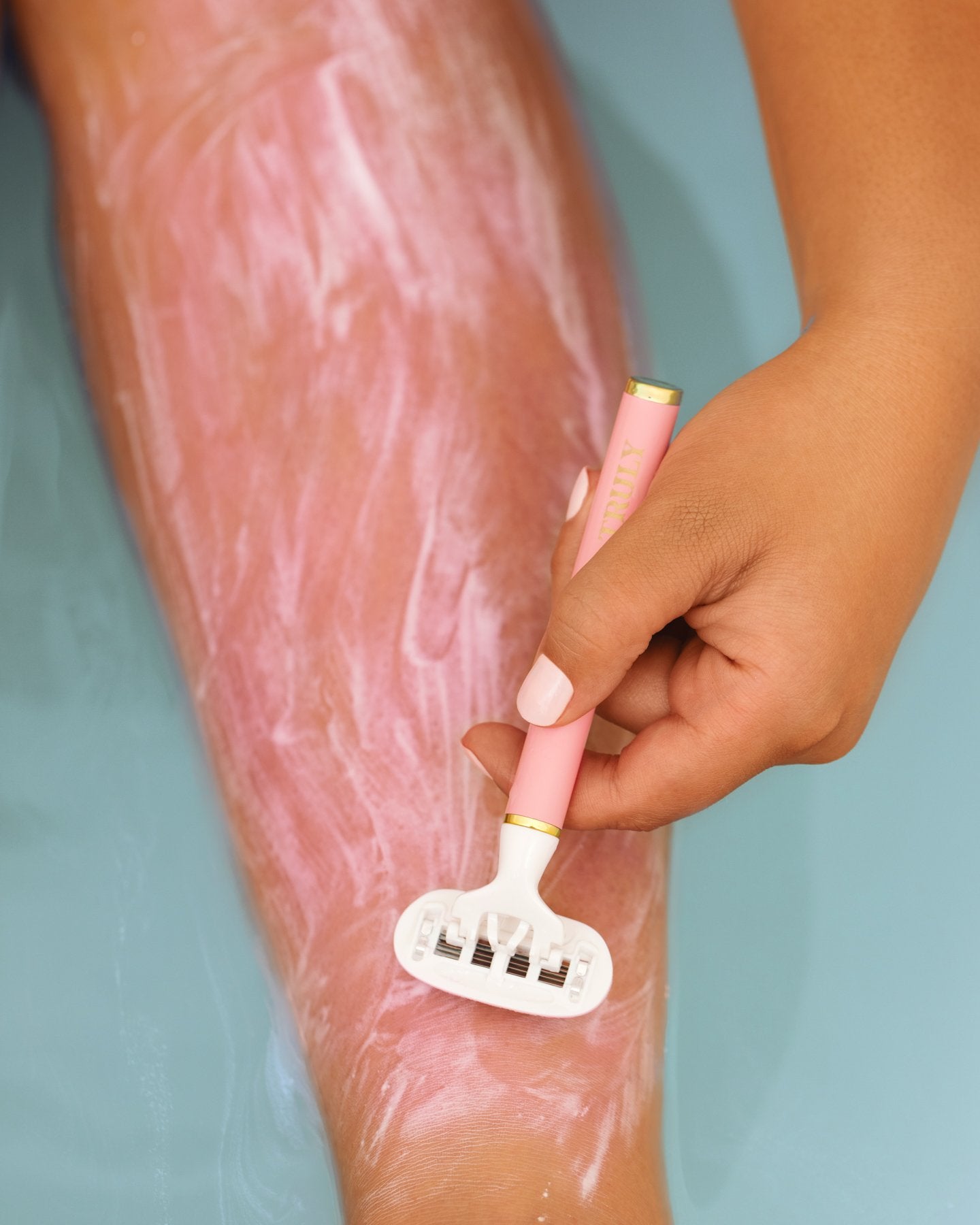 A close-up shows a hand shaving a lower leg with the Truly Beauty Life Shaver Starter Razor and soothing aloe shaving cream for sensitive skin, all over a blue surface that appears to be a bathtub.